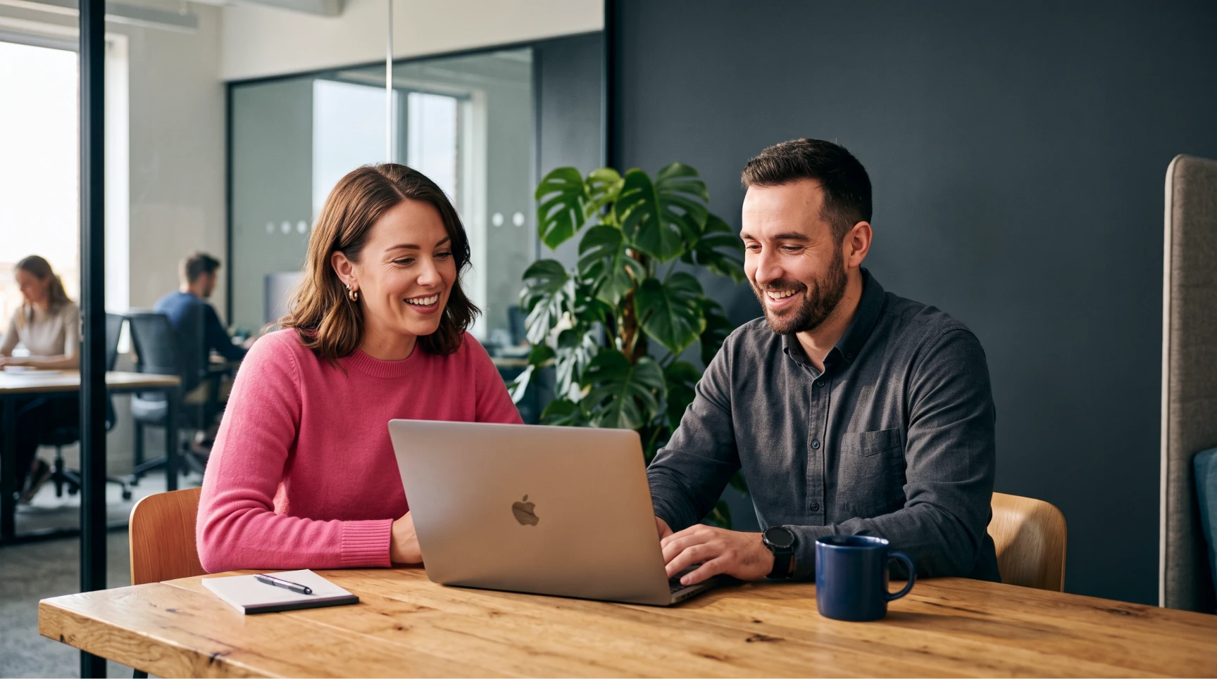 Um homem e uma mulher sorriem enquanto olham para a tela de um notebook sobre uma mesa de madeira, realizando o sourcing de candidatos durante uma etapa estratégica de recrutamento e seleção.
