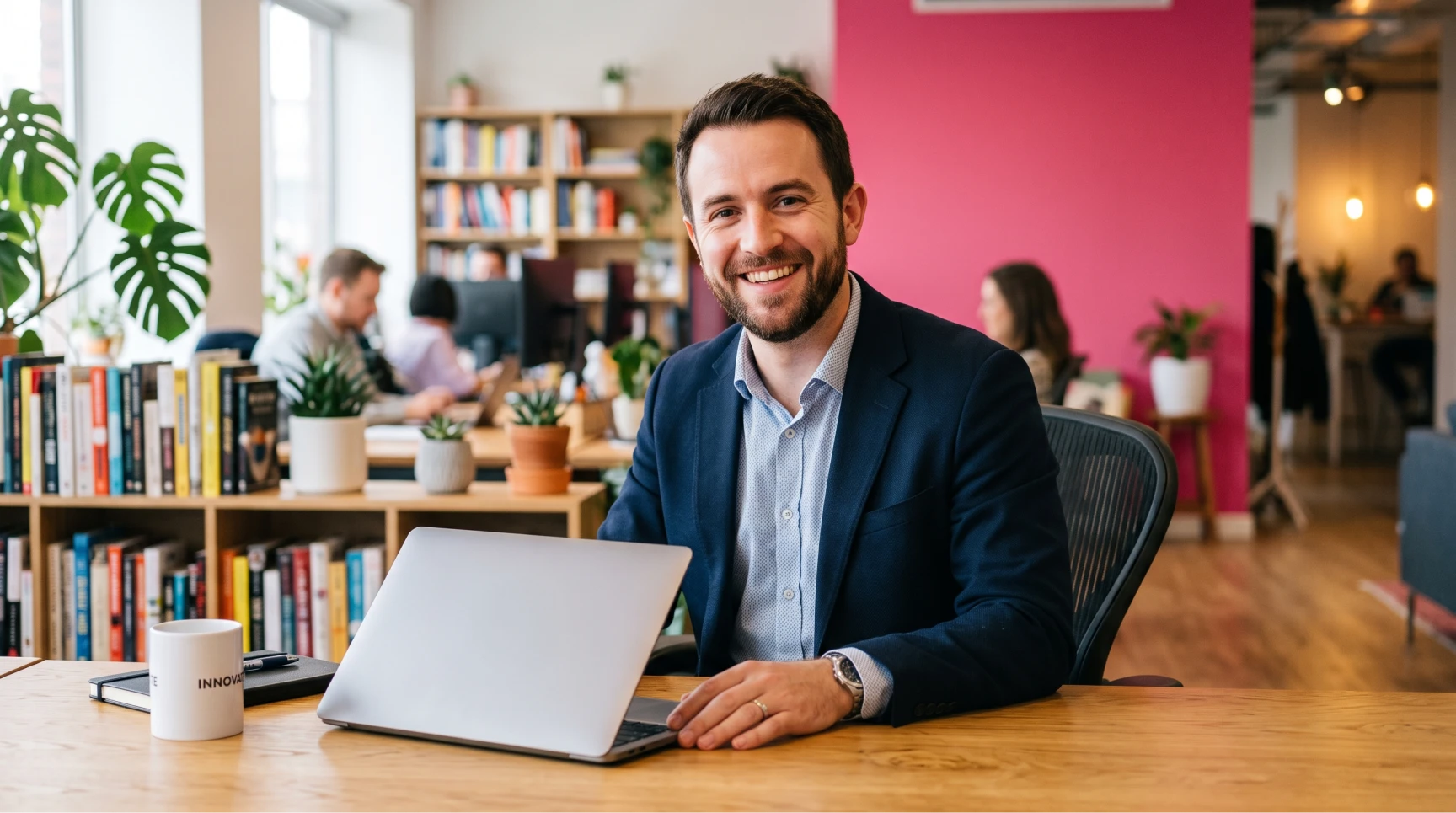 Um homem barbudo e sorridente vestindo blazer azul escuro senta-se atrás de sua mesa com um laptop aberto, representando a figura de um headhunter tecnologia atuando como parceiro de recrutamento de empresas de software.