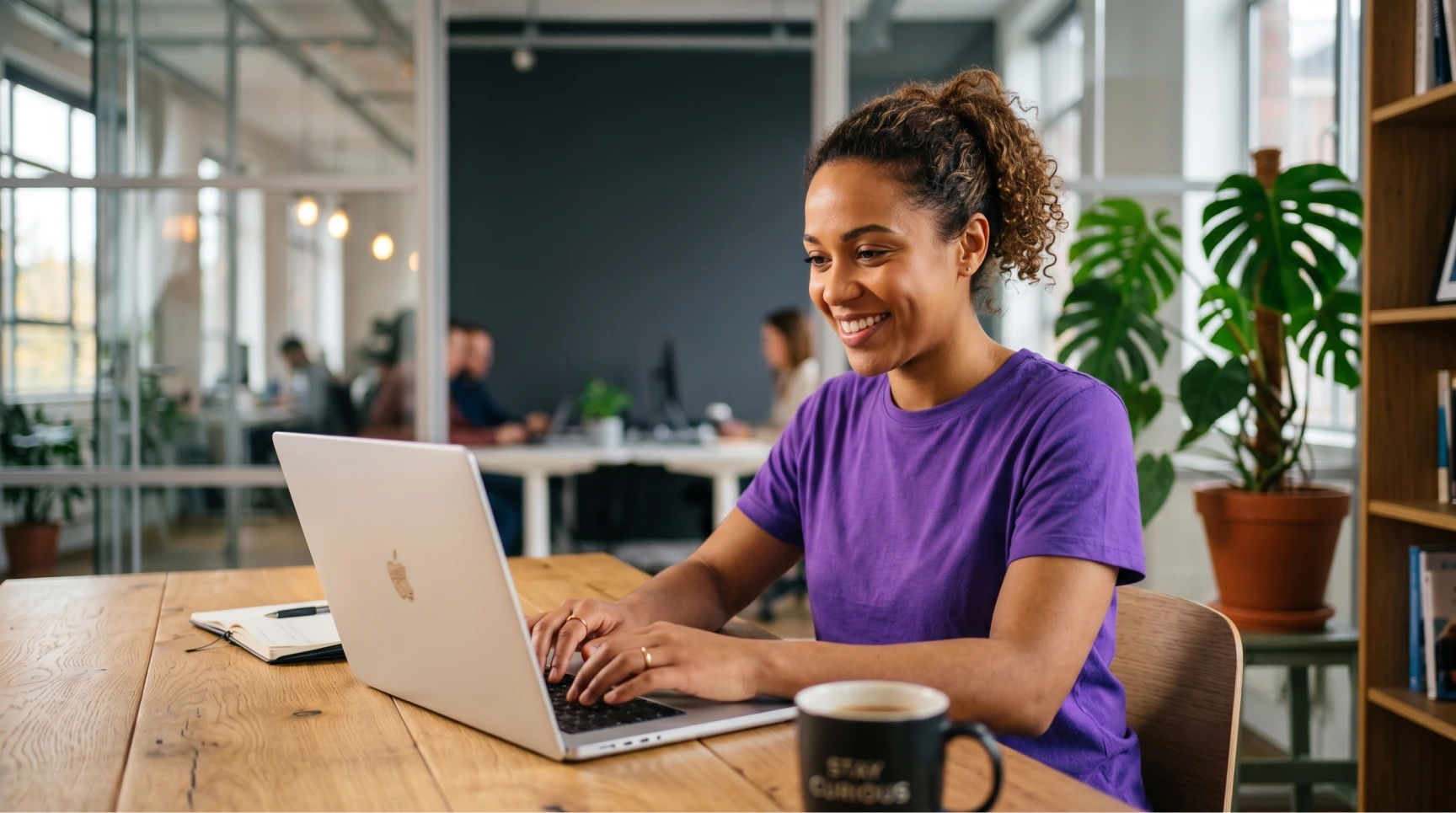 Mulher de cabelos cacheados sorrindo enquanto digita em seu notebook em um escritório moderno, ilustrando o trabalho diário de recrutamento remoto para o preenchimento de vagas tech.